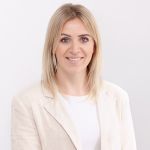 Eine Frau mit langen blonden Haaren lächelt in die Kamera. Sie trägt einen hellen Blazer über einem weißen Oberteil. Der Hintergrund ist schlicht weiß und vermittelt ein sauberes und professionelles Erscheinungsbild.