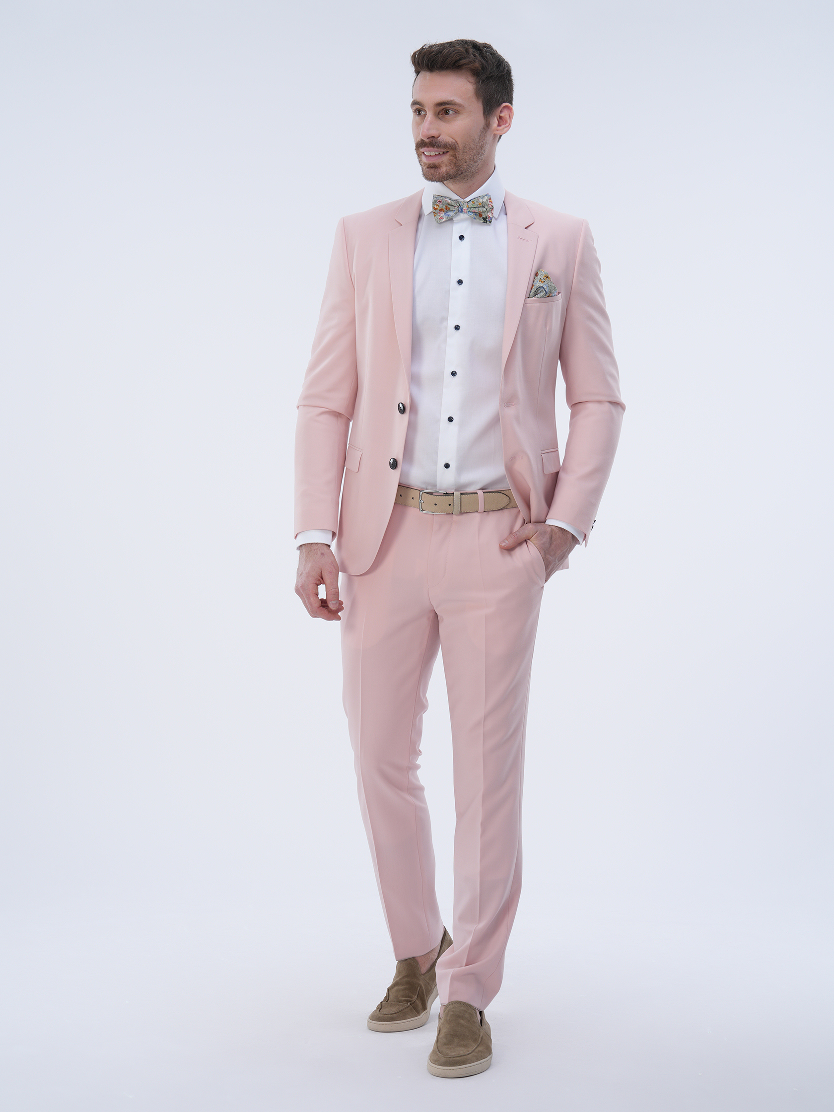 Ein Mann steht in einem rosa Anzug mit weißem Hemd und gemusterter Fliege. Er hat die rechte Hand in der Tasche und trägt braune Schuhe. Der Hintergrund ist schlicht weiß.