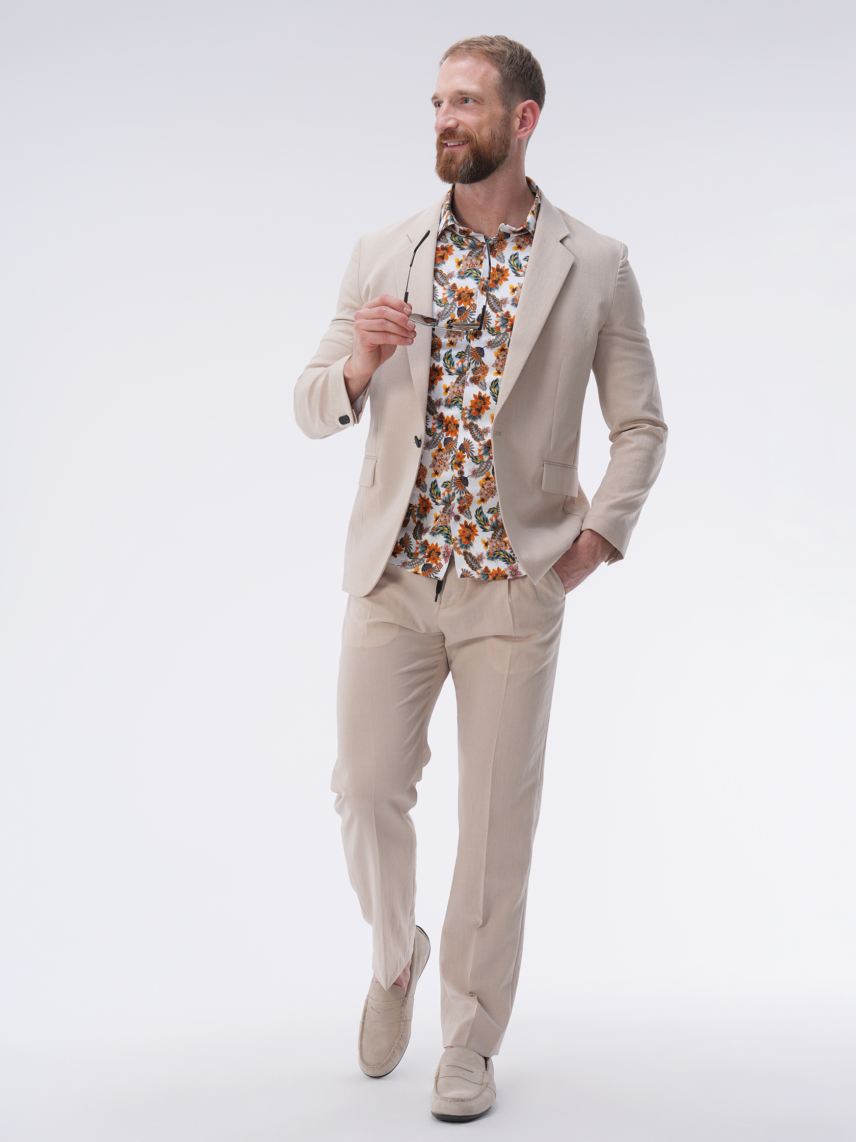 Ein Mann in einem hellbeigen Anzug und einem bunten Hemd mit Blumenmuster hält eine Sonnenbrille in der Hand, steht lächelnd vor einem schlichten weißen Hintergrund. Er trägt Freizeitschuhe und strahlt eine entspannte und fröhliche Ausstrahlung aus.