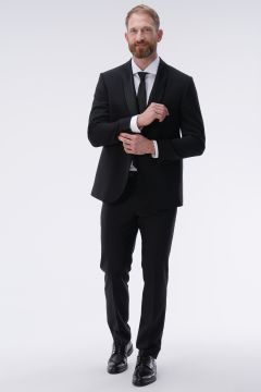Ein Mann in schwarzem Anzug, weißem Hemd und schwarzer Krawatte richtet lächelnd seinen linken Ärmel. Er steht vor einem schlichten weißen Hintergrund und trägt schwarze Schuhe. Sein Haar und sein Bart sind hellbraun.