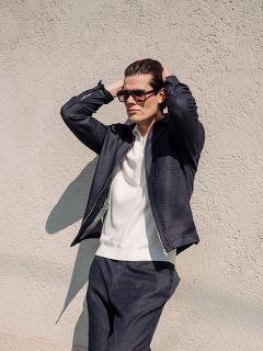 Ein Mann, der eine Sonnenbrille, eine marineblaue Jacke und eine dazu passende Hose trägt, steht mit den Händen auf dem Kopf vor einer hellen, strukturierten Wand.
