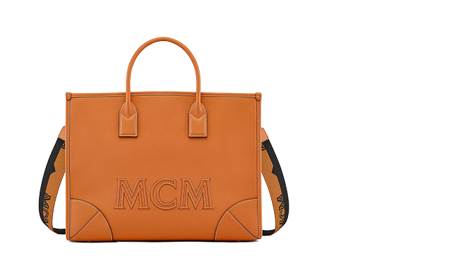 Orangefarbene MCM-Handtasche mit zwei Griffen und einem abnehmbaren schwarzen Riemen mit dem Markennamen. Die Tasche hat ein strukturiertes Design mit sichtbaren Nähten und Eckdetails und trägt auf der Vorderseite deutlich die Prägung „MCM“.