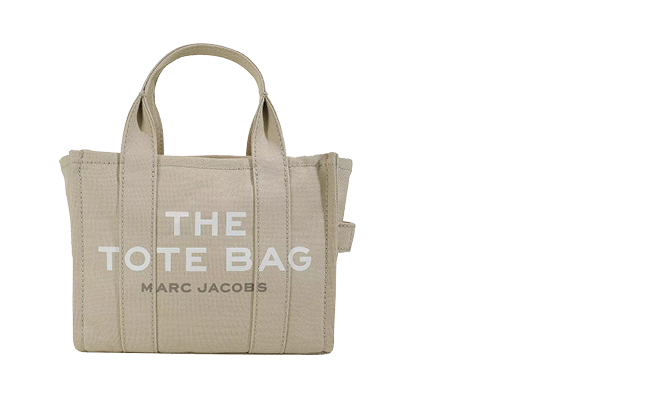 Beigefarbene Tragetasche mit Griffen und den Worten „THE TOTE BAG“ und „MARC JACOBS“ in Weiß auf der Vorderseite aufgedruckt. Die Tasche ist schlicht und strukturiert, mit sichtbaren Nähten und einem seitlichen Riemendetail.