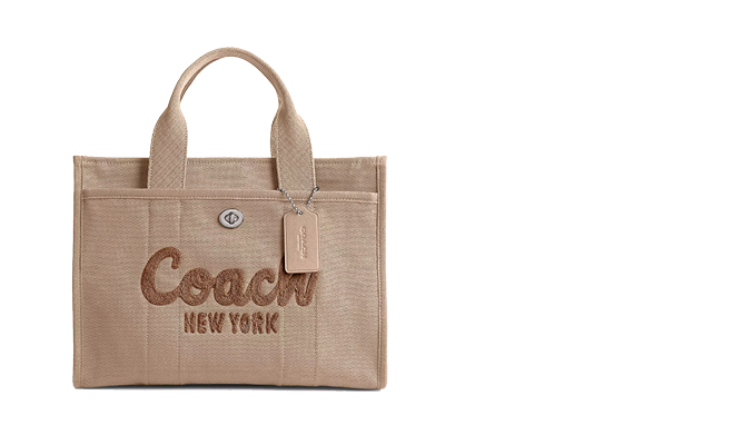 Beigefarbene Coach-Tragetasche mit aufgesticktem „Coach New York“ auf der Vorderseite. Die Tasche hat zwei kurze Griffe und ein daran befestigtes Gepäcketikett. Der Hintergrund ist weiß.