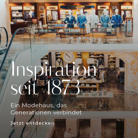 Ein mehrstöckiger Innenbereich eines Modegeschäfts mit eleganten Kleiderpräsentationen und einer zentralen Rolltreppe. Das Bild enthält den Text in deutscher Sprache: „Inspiration seit 1873. Ein Modehaus, das Generationen verbindet. Jetzt entdecken.“.