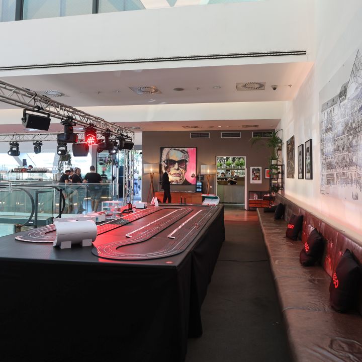 Raum mit einem großen Slotcar-Renntisch in der Mitte. Gepolsterte Sitzgelegenheiten säumen eine Wand und ein Wandgemälde mit einer Stadtlandschaft schmückt eine andere. Deckenleuchten sorgen für eine gemütliche Atmosphäre und an der Rückwand hängt ein gerahmtes Poster.