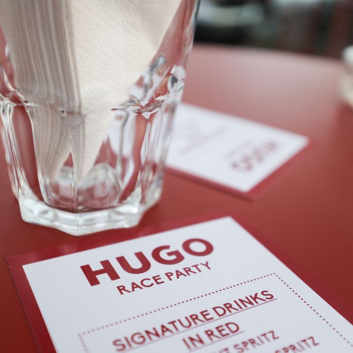Nahaufnahme eines gedeckten Restauranttisches mit einem Glas, in dem gefaltete Servietten liegen. Auf dem Tisch liegt eine Menükarte mit der Überschrift „Hugo Race Party“ und einer Liste „Signature Drinks“, darunter „Vermut Spritz“ und „Crodino Rosso Spritz“.