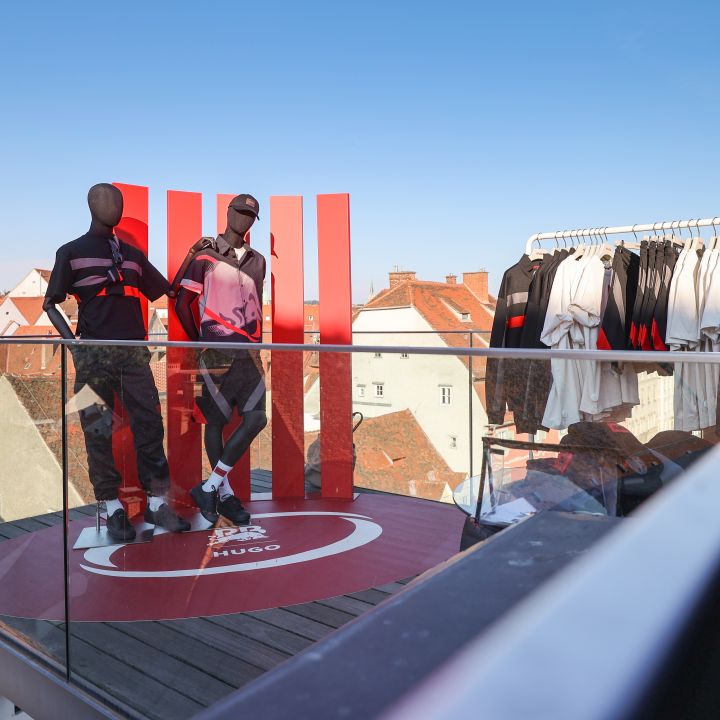 Zwei Schaufensterpuppen in sportlicher Kleidung stehen auf einem Dachdisplay mit Glasgeländer. Rechts ist ein Ständer mit ähnlicher Kleidung. Im Hintergrund sind Dächer und ein klarer blauer Himmel zu sehen.