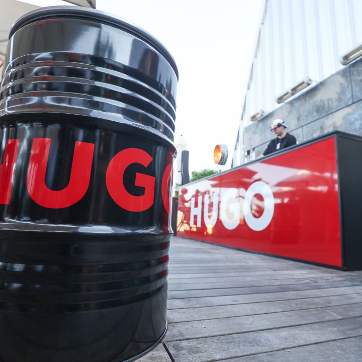 Schwarzes Metallfass mit der Aufschrift „HUGO“ in roten Buchstaben auf einem Holzdeck; ein DJ steht hinter einem rot-schwarzen Pult, ebenfalls mit der Aufschrift „HUGO“. Im Hintergrund sind Menschen unter einem Vordach bei einer Veranstaltung im Freien zu sehen.