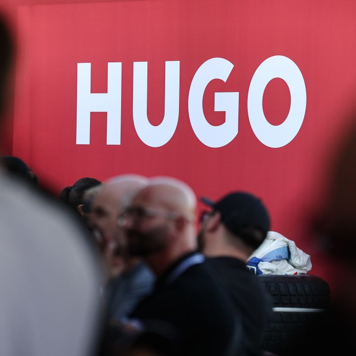 Ein großes rotes Schild mit dem Wort „HUGO“ in fetten weißen Buchstaben. Im Vordergrund sind verschwommene Figuren von Menschen zu sehen, die unscharf stehen oder gehen.