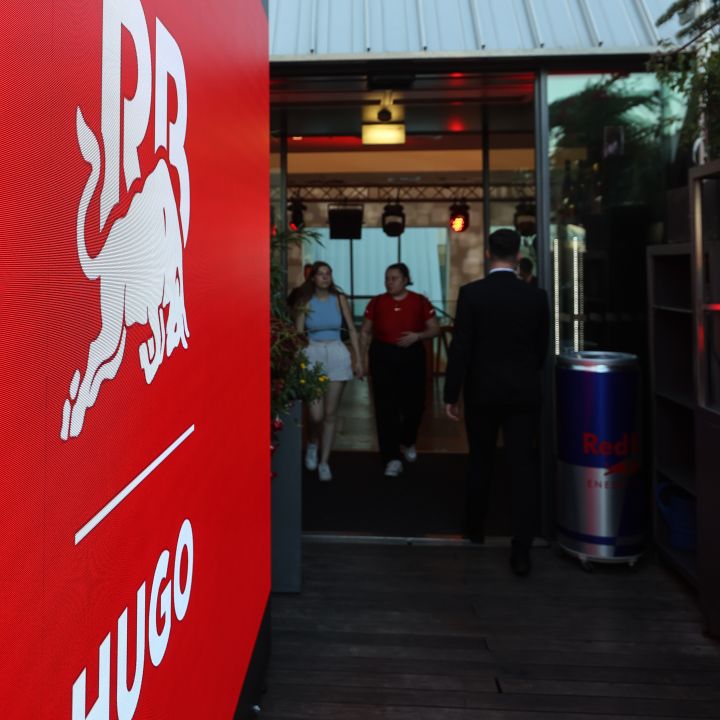 Großes rotes Schild mit dem Logo von Hugo Red Bull auf der linken Seite. Zwei Personen gehen in ein modernes Gebäude im Hintergrund. Rechts steht ein Mann im Anzug und in der Nähe ein Heizgerät mit der Marke Red Bull.