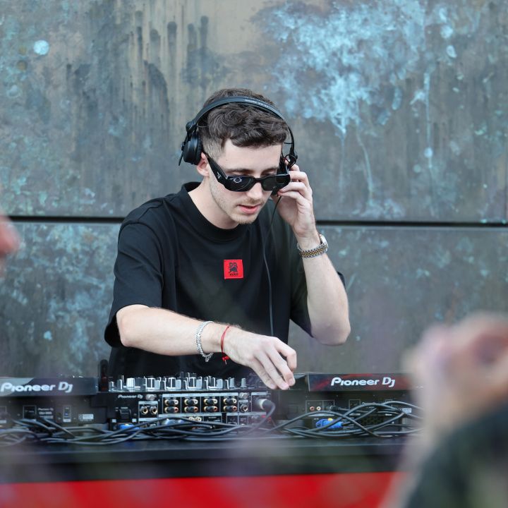 Ein DJ mit Sonnenbrille und Kopfhörern ist konzentriert damit beschäftigt, die Regler eines Pioneer DJ-Mixers einzustellen. Er spielt Musik im Freien, mit verschwommenen Menschen im Vordergrund und einer strukturierten Wand im Hintergrund.
