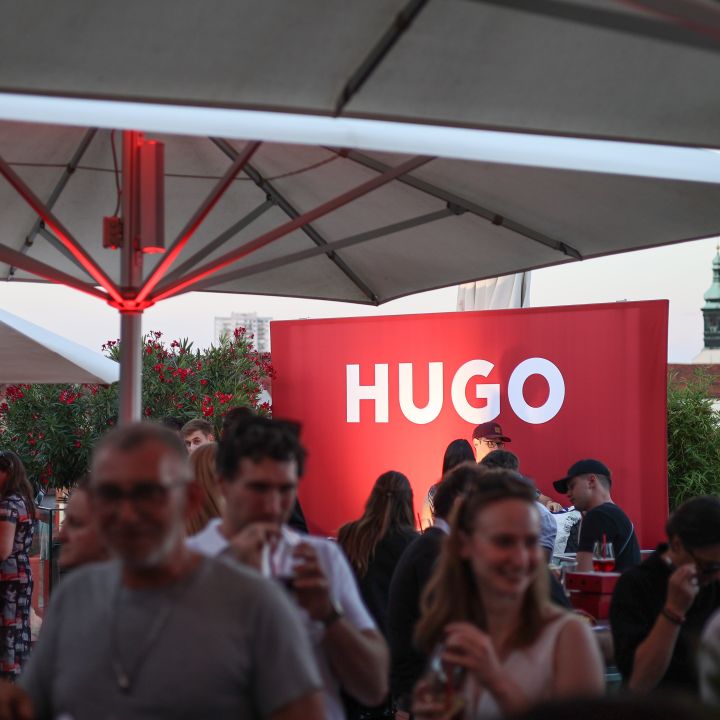 Eine lebhafte Veranstaltung im Freien, bei der Menschen unter großen Sonnenschirmen gesellig zusammensitzen. Auffällig ist ein rotes Schild mit der Aufschrift „HUGO“ in fetten weißen Buchstaben. Im Hintergrund sind ein klarer Himmel und entfernte architektonische Strukturen zu sehen.