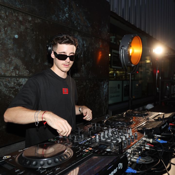 Ein DJ mit Sonnenbrille und schwarzem Hemd tritt bei einer Veranstaltung im Freien auf. Er verwendet eine Konsole mit verschiedenen Bedienelementen und Tasten. Im Hintergrund sind eine strukturierte Wand und ein rundes Licht zu sehen, dessen Strahl den Bereich beleuchtet.