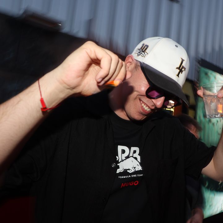 Eine Person mit schwarzem Hemd, Sonnenbrille und weißer Kappe lächelt und tanzt, während sie in einer Hand ein Getränk hält. Der Hintergrund ist ein verschwommenes Wandgemälde auf einer Metallstruktur, das eine Außenkulisse suggeriert.