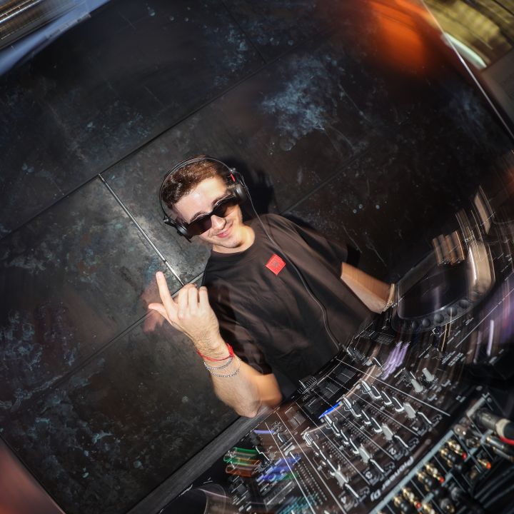 Ein DJ mit Sonnenbrille und Kopfhörern mixt Songs an einem Plattenteller in einem schwach beleuchteten Club. Bunte Lichter verschwimmen und erzeugen eine dynamische und energiegeladene Atmosphäre.
