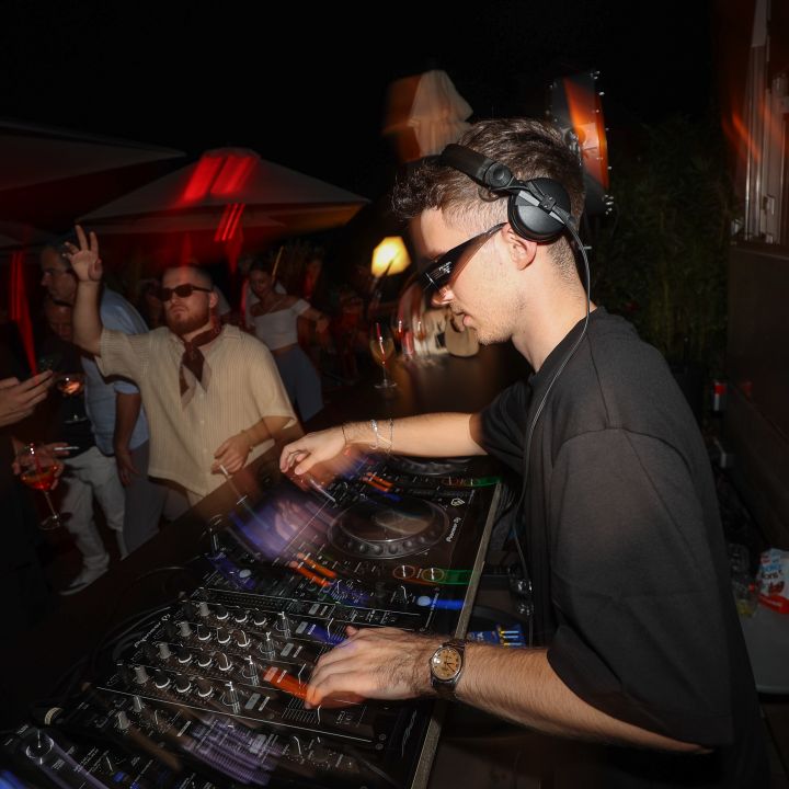 Ein DJ mit Sonnenbrille und Kopfhörern mixt Musik auf einem Mischpult bei einer lebhaften Veranstaltung im Freien. Zwei Personen im Hintergrund genießen die Atmosphäre, eine hält ein Getränk in der Hand. Bunte Lichter und ein dunkler Nachthimmel sorgen für eine lebhafte Partyszene.