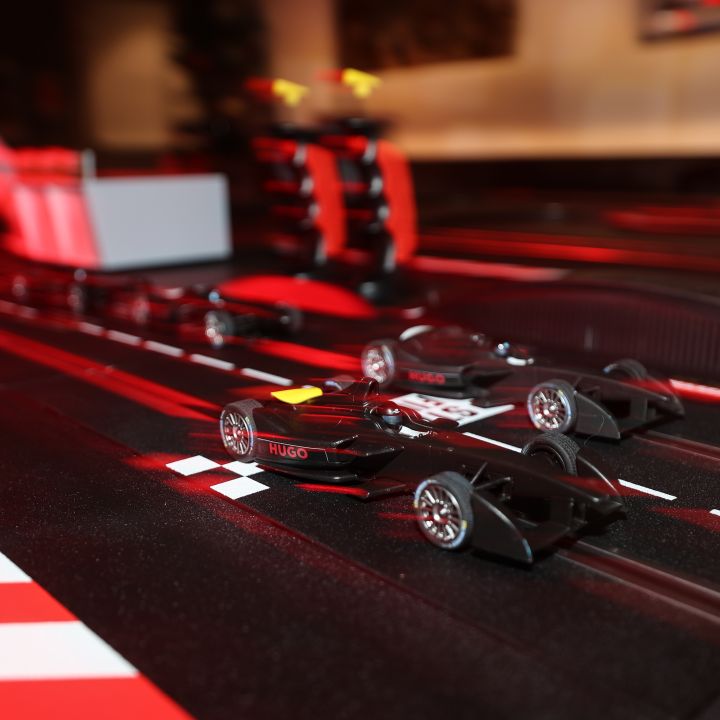 Nahaufnahme von zwei Spielzeug-Rennautos auf einer schwarzen Strecke mit roten und weißen Markierungen. Die Szene enthält rote und schwarze Barrieren und Strukturen, die eine Miniatur-Rennumgebung schaffen.