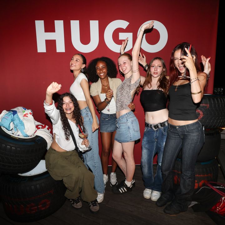 Eine Gruppe von sechs jungen Frauen posiert fröhlich vor einem roten Hintergrund mit dem Text „HUGO“. Sie sind leger gekleidet und stehen zwischen gestapelten Reifen, wobei einige verspielte Handbewegungen machen. Die Szene wirkt festlich und energiegeladen.