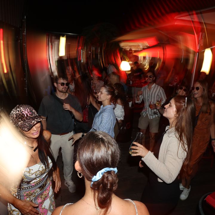 Ein lebendiges, verschwommenes Bild von Menschen, die auf einer Party mit wirbelnden Lichtern und einem warmen Farbschema tanzen. Die Personen, in lässiger Sommerkleidung gekleidet, lächeln und halten Getränke, wodurch eine Atmosphäre der Freude und Bewegung entsteht.