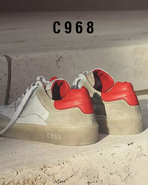 Ein Paar stylische Sneaker auf einer Steinoberfläche. Sie haben weiße und graue Oberteile mit kräftigen roten Akzenten an der Ferse. Die Nummer „C968“ erscheint auf der Stufe im Hintergrund und auf den Schuhsohlen.