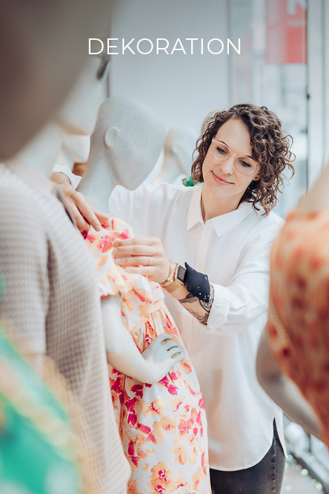 Eine Frau mit lockigem Haar, Brille und weißem Hemd richtet ein buntes Kleid an einer Schaufensterpuppe. Die Szene scheint ein Bekleidungsgeschäft mit weiteren Schaufensterpuppen zu sein. Oben im Bild ist das Wort „DEKORATION“ zu sehen.