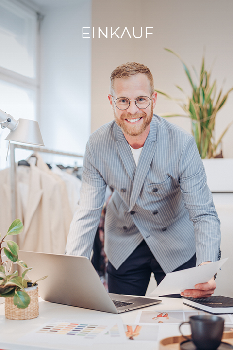 Lächelnder Mann mit Brille und Bart lehnt über einem Schreibtisch und hält Papiere in der Hand. Er trägt einen gestreiften Blazer und steht in einem modernen Büro mit einer Pflanze und einem Laptop. Kleiderständer und Farbmuster sind zu sehen. Über ihm steht das Wort „EINKAUF“.
