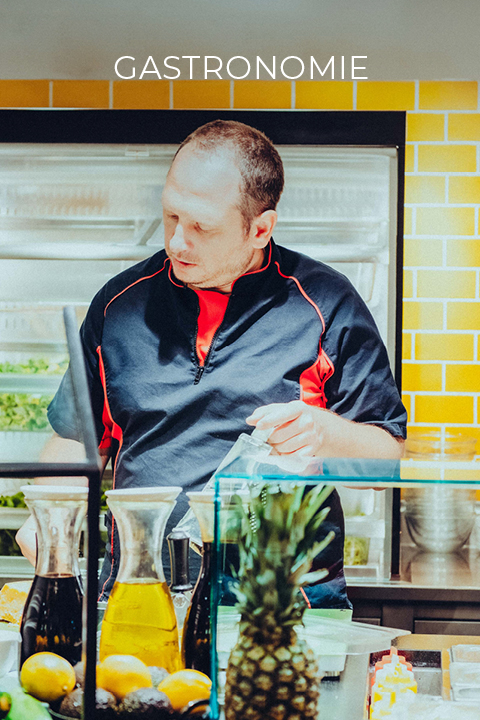 Hinter einem Tresen mit gelben Kacheln arbeitet ein Mann in schwarz-roter Uniform. Vor ihm stehen Karaffen mit Flüssigkeiten, eine Ananas und andere Früchte. Darüber steht das Wort „Gastronomie“.