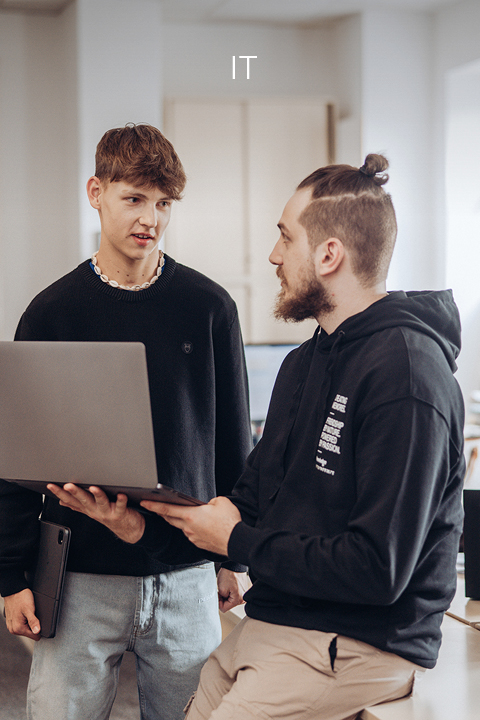 Zwei Männer stehen in einem Büro. Einer steht und hält einen Laptop in der Hand, der andere sitzt und unterhält sich. Beide sind leger gekleidet. Das Wort „IT“ steht oben im Bild und weist auf einen IT-Kontext hin.