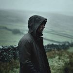 Eine Person, die einen dunklen Regenmantel mit Kapuze trägt, steht in einer nebligen, ländlichen Landschaft mit Steinmauern und sanften Hügeln im Hintergrund. Die Szene wirkt kalt und bewölkt.