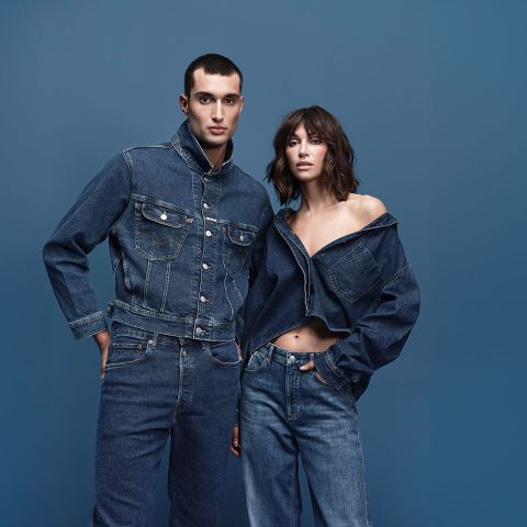 Ein Mann und eine Frau posieren vor einem blauen Hintergrund, beide tragen Jeanskleidung. Der Mann trägt eine Jeansjacke und Jeans, während die Frau eine schulterfreie Jeansjacke und Jeans trägt.