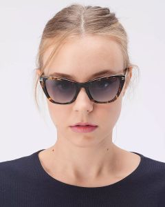 Eine Person mit zurückgebundenem hellbraunem Haar, einer Schildpatt-Cat-Eye-Sonnenbrille und einem dunklen Oberteil blickt direkt in die Kamera. Der Hintergrund ist schlicht weiß.