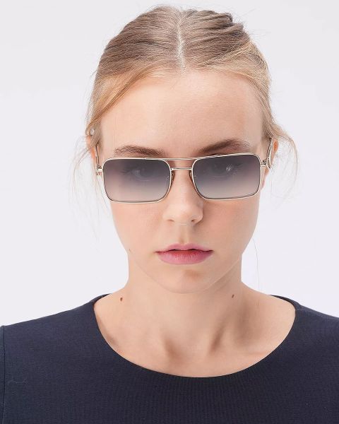 Eine Person mit hellbraunem Haar trägt eine rechteckige Sonnenbrille und ein marineblaues Oberteil und steht vor einem schlichten weißen Hintergrund.