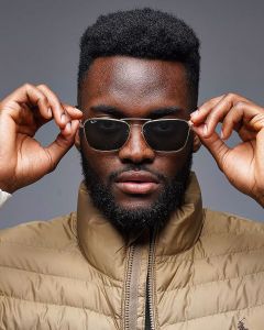 Eine Person mit Sonnenbrille rückt sie mit beiden Händen zurecht. Sie hat einen Bart und trägt eine hellbraune Pufferjacke. Der Hintergrund ist neutral grau.