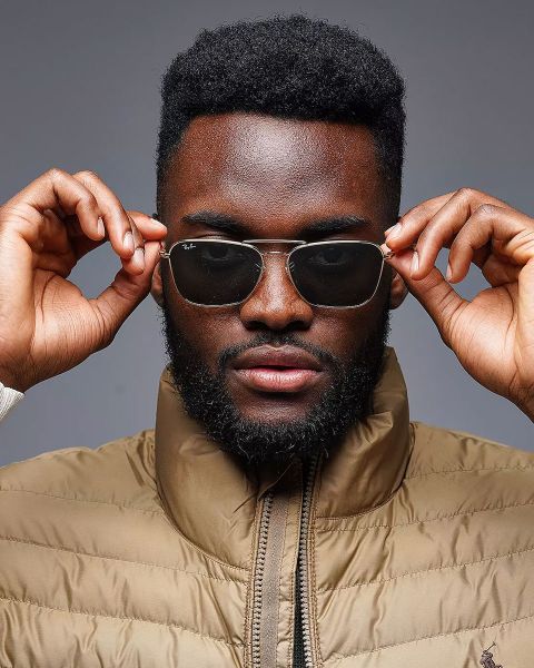 Eine Person mit Sonnenbrille rückt sie mit beiden Händen zurecht. Sie hat einen Bart und trägt eine hellbraune Pufferjacke. Der Hintergrund ist neutral grau.