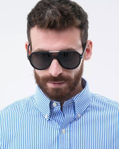 Ein bärtiger Mann trägt eine dunkle Sonnenbrille und ein blau-weiß gestreiftes Hemd mit zugeknöpftem Kragen. Er blickt leicht nach unten. Der Hintergrund ist schlicht weiß.