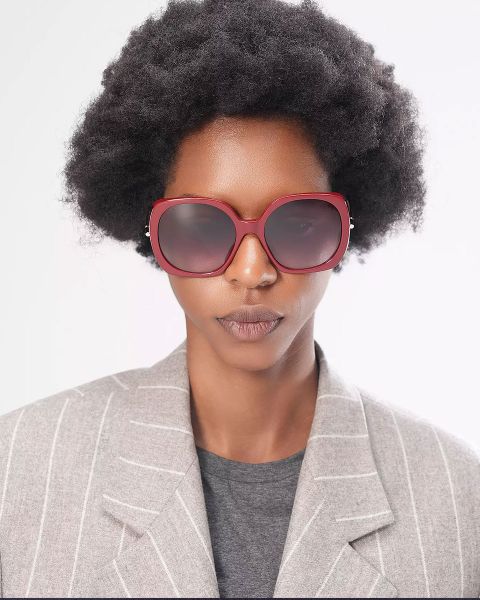 Eine Person mit mittellanger Afrofrisur trägt eine große, runde rote Sonnenbrille und einen grauen Blazer über einem dunklen Hemd. Der Hintergrund ist schlicht weiß.