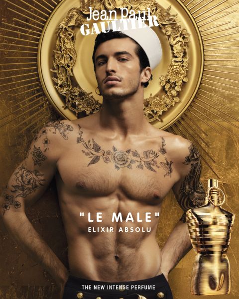 Ein hemdloses männliches Model mit Tattoos trägt eine Matrosenmütze vor einem goldenen Hintergrund und wirbt für Jean Paul Gaultiers Parfüm „Le Male“ Elixir Absolu. In der unteren rechten Ecke befindet sich eine goldene Parfümflasche. Der Text lautet: „Das neue intensive Parfüm.