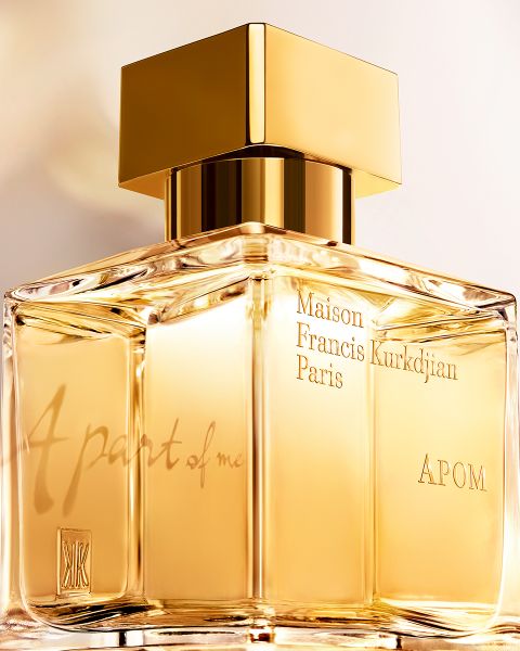 Eine Nahaufnahme einer rechteckigen Parfümflasche aus Glas mit goldenen Akzenten. Auf dem Etikett steht „Maison Francis Kurkdjian Paris, APOM“. Die Flasche ist mit einer goldenen Flüssigkeit gefüllt und an der Seite sind die Worte „Ein Teil von mir“ eingraviert.