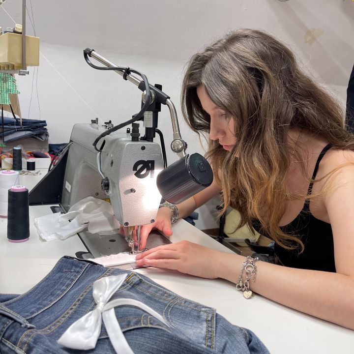 Eine Frau mit langen Haaren näht konzentriert an einer Maschine in einem gut beleuchteten Raum. Im Vordergrund ist eine Jeans mit einer weißen Schleife zu sehen. Im Hintergrund sind Regale mit Nähmaterial zu sehen.
