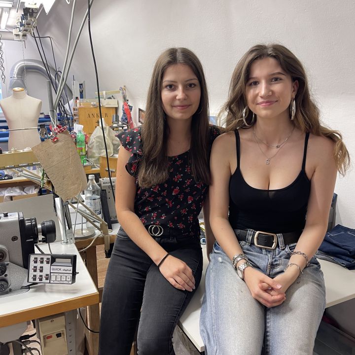 Zwei Frauen sitzen in einer Nähwerkstatt. Sie lächeln und schauen in die Kamera, umgeben von Nähmaschinen, Stoffen und Bastelutensilien. Eine trägt ein geblümtes Top und schwarze Jeans, die andere ein schwarzes Tanktop und helle Jeans.