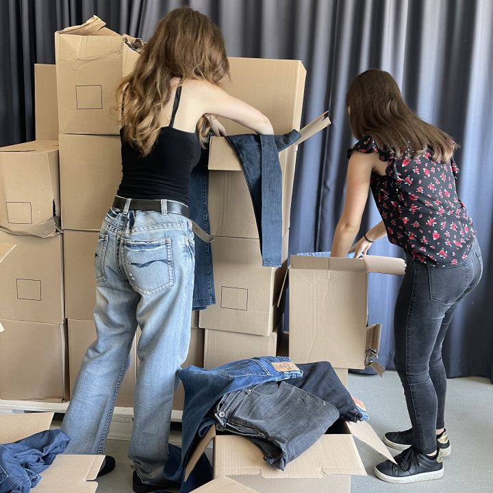 Zwei Frauen sortieren Kartons voller Jeans. Sie stehen neben einem Stapel Kartons und ziehen die Jeans heraus und sortieren sie. Eine Frau trägt ein schwarzes Oberteil und helle Jeans, die andere ein geblümtes Oberteil und dunkle Jeans, im Hintergrund ein Vorhang.