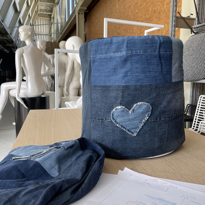 Ein Aufbewahrungskorb aus Jeansstoff mit einem Patchwork-Herzmuster steht auf einem Tisch neben einem gefalteten Jeanskleidungsstück. Im Hintergrund sind Schaufensterpuppen und Regale sowie Skizzen auf Papier zu sehen.