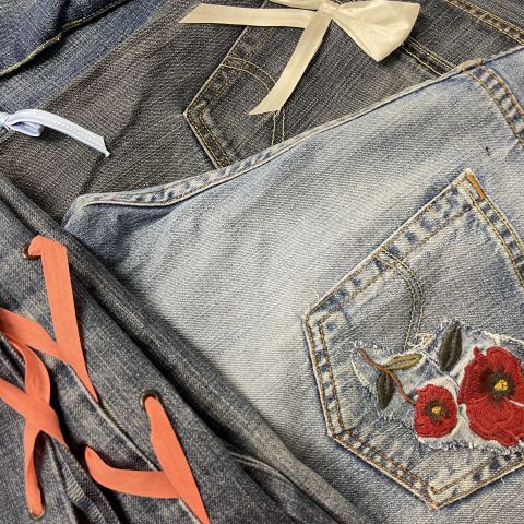 Nahaufnahme von Jeanskleidung mit dekorativen Elementen. Dazu gehören eine weiße Schleife am Hosenbund, rote Blumen, die auf eine Tasche gestickt sind, und rote Schnürdetails entlang einer Naht. Die Jeans variieren im Farbton von Hell- bis Dunkelblau.