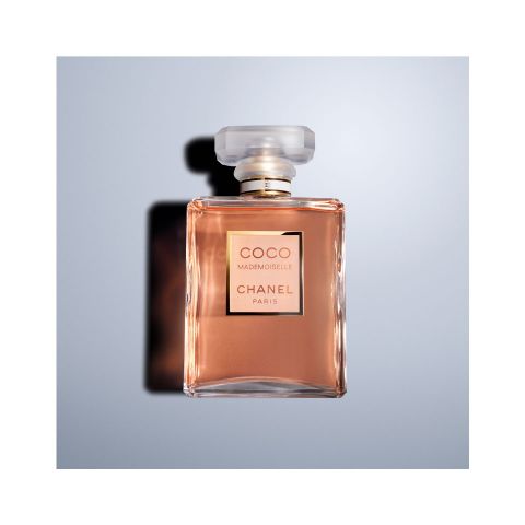 Eine quadratische Glasflasche mit Coco Mademoiselle Chanel Paris-Parfüm mit einer goldbraunen Flüssigkeit im Inneren. Die Flasche hat einen facettierten, klaren Deckel und einen silbernen Hals. Sie ist vor einem hellblauen Hintergrund fotografiert.