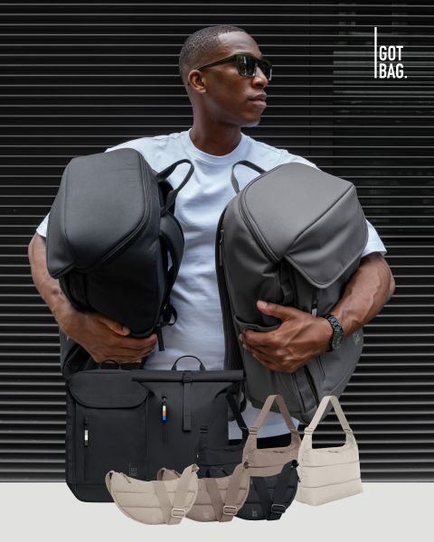 Mann mit Sonnenbrille hält große graue Taschen vor einem dunkel gestreiften Hintergrund. Unter ihm sind verschiedene Taschen in verschiedenen Größen und Farben, alle ordentlich ausgestellt. Das Logo oben rechts lautet „GOT BAG“.