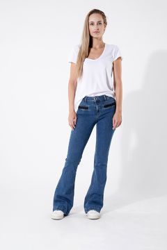 Eine Person in einem weißen T-Shirt und blauen Schlagjeans steht vor einem schlichten weißen Hintergrund. Sie hat lange Haare und trägt weiße Turnschuhe. Sie posiert mit einer Hand in der Tasche und der anderen auf der Hüfte.