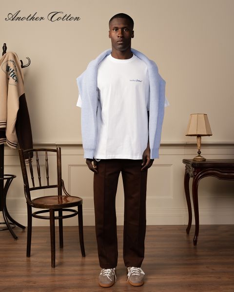 Eine Person steht in einem Raum, trägt einen hellblauen Pullover über den Schultern, ein weißes T-Shirt und braune Hosen. Im Raum stehen ein Holzstuhl, eine Vintage-Stehlampe und eine Jacke an einer Garderobe. Auf dem Bild ist der Text „Another Cotton“ zu sehen.