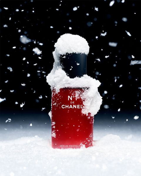 Eine rote Flasche Serum N°1 DE CHANEL, teilweise mit Schnee bedeckt, steht aufrecht vor einem dunklen Hintergrund. Schneeflocken fallen herab und verleihen dem Produkt eine winterliche Atmosphäre.