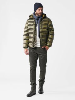 Ein Mann in einer grünen Pufferjacke, grünen Hosen, schwarzen Stiefeln, einer marineblauen Mütze und einem grauen Schal steht vor einem schlichten weißen Hintergrund. Er blickt mit entspannter Haltung zur Seite und hat die Hände in den Taschen.